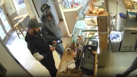 CCTV screencap of two men in a coffee shop. One is holding the chip and pin machine. The other is standing at the counter looking at the employee serving at the till. 