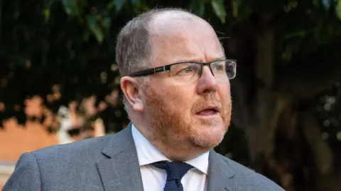 George Freeman is a middle aged man, with a balding head and red facial hair.