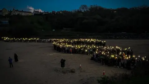 BBC a group of people on a beach, each holding a light so that the group makes an s shape