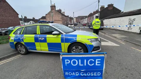 Kate Bradbrook/BBC A marked blue and yellow fluorescent police car and police officer stood behind a police cordon area on Newcomen Road in Wellingborough. A blue sign with white writing says POLICE ROAD CLOSED.