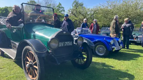 Four classic cars, three green and one blue, are on grass, with hedges in the background. Two people are sitting in one of the green cars in the foreground on the left and others are standing up.