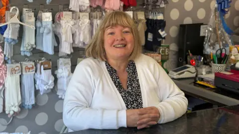 A woman smiles from behind shop counter 