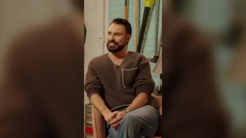 Rylan Clark has black hair and is wearing a brown jumper and grey trousers. He sits crossed-legged and smiles.