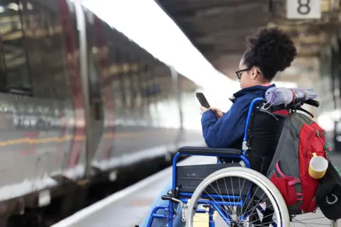 Getty Images Pengguna kursi roda wanita menunggu di platform kereta api, menggunakan teleponnya.