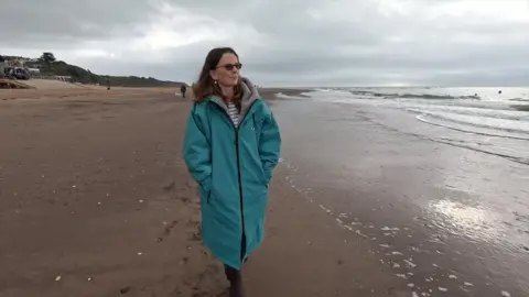 BBC A woman wearing a long blue jacket walking along the beach with the sea to her right.
