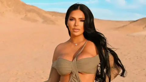 klaudiaglam Klaudia Zakrzewska in a desert with blue sky. She wears an off-the-shoulder bikini style top which is taupe. Her hair is long and dark and behind her shoulders. She looks directly at the camera and has a small pendant necklace on. 