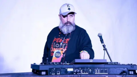 Konrad Marciak A person wearing a baseball cap and a dark sweatshirt stands behind professional DJ equipment, with one hand on the controls. A microphone on a stand sits to the right, and the scene is lit by bright white and blue-tinted stage lighting against a simple backdrop.