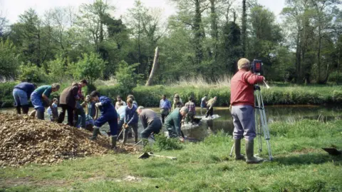 Owen Sennitt/BBC A man with a video camera on a tripod stands looking towards a group of people creating a riffle, which is a gravel river bed, along the River Wensum in the early 1990s.