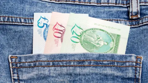 Getty Images Three bank notes in the pocket of denim jeans. The £5 note is blue, the £10 note is red and the £20 note is green.