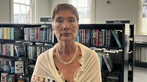 Aine Andrews has short brown hair and is wearing a white shawl and a white necklace. She is standing in the middle of Falls Road Library, black bookcases full of books can be seen behind her.