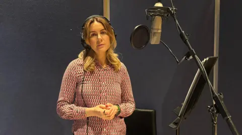 Olivia Graham stands in a recording studio in front of a mic, wearing headphones. She is wearing a geometric pattern pink and blue blouse and has her blonde/brown hair in curls around her shoulders