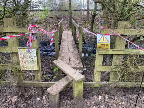 Ben Thornely A style and wooden footbridge over the ditch with red tape draped across it