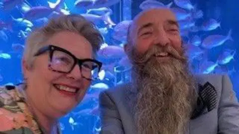 John Gifford and Lisa Gifford pictured smiling in a blue lit room. John has a long grey beard and a moustache which curls at the ends, he wears a blue suit jacket with a dark blue pocket square while Lisa has a grey pixie cute, wears red lipstick, large black framed glasses and a pink, yellow and blue floral top.