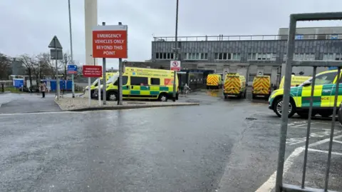 BBC Ambulances parked outside Derriford Hospital. A large red sign is leading into the car park and says emergency vehicles only beyond this point.