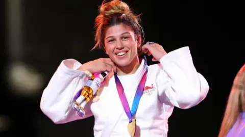Getty Images Acelya has a medal around her neck and is holding a small mascot in her hand as she smiles at the camera after winning. Her brown hair it tied up on her head.