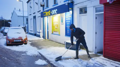 PA Media A man clears snow from a pavement in Dowlais, Merthyr Tydfil after. Storm Goretti. He's dressed in a hooded coat, a car nearby is covered in snow