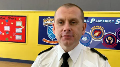 Supt John Wilson is in his police uniform, shirt and tie, standing in a school corridor. The wall behind him is yellow and has several posters and boards.