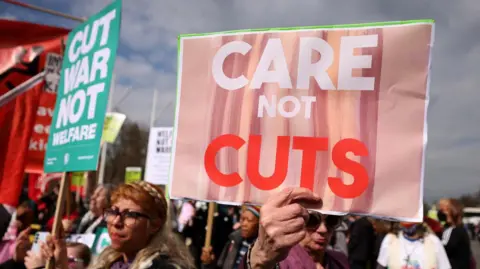 A crowd of people protesting, holding up signs including "care not cuts" and "cut war not welfare". 