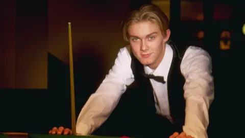 Getty Images A young Paul Hunter poses for a photo as he leans against a snooker table. A cue stands by his right hand. He has floppy blond hair and is wearing a bow tie, white shirt and waistcoat.