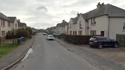 A street with two-storey residential buildings on either sides. There are four cars parked on driveways and on the road.