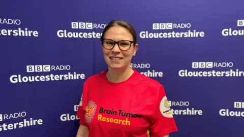 A woman with dark brown hair which has been tied back in a loose ponytail smiles as she stands in front of a purple BBC Radio Gloucestershire background. She is wearing black-rimmed glasses and a bright red Brain Tumour Research top.
