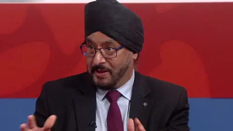 A man in a black turban, purple suit and black glasses talks in a TV studio.