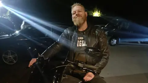 Handout Paul Hart sits on a motorbike at night. He is wearing a black top and leathers and a bright light is shining down. There are cars parked nearby.