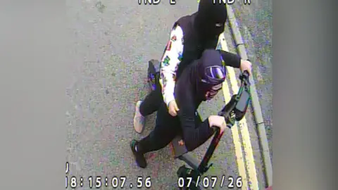 CCTV image of two people on an electric scooter. They have their heads and faces covered and are wearing mostly black clothing