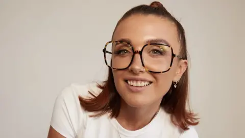 Candid Studios Ellie is looking straight at the camera. She is wearing big square tortoise shell glasses. Her hair is shoulder length with a reddish tint and is slightly pulled back. She has a silver ring piercing in her nose and three silver hoops in her ears. She's wearing a white t-shirt.