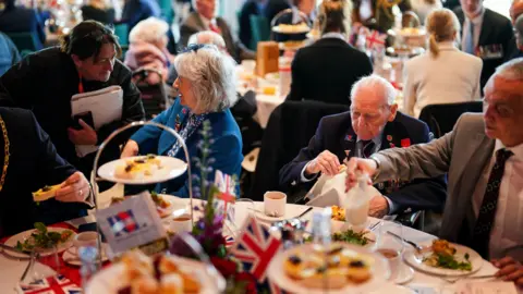 PA Media People sit around a table with cups of tea and plates of food.