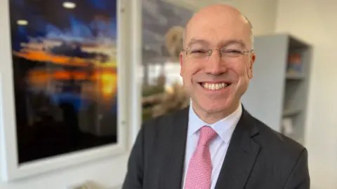 Stuart Woodward/BBC Jonathan Denby from Greater Anglia, smiling at the camera, wears a dark grey suit, light blue shirt and pink patterned tie, and wears rimless glasses. He is stood in front of two examples of artwork, one featuring a dog and the other featuring a sunset over a beach.