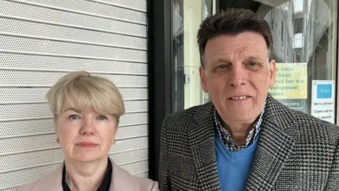 Lara King A man and a woman stand in front of shops with shutters down. The woman has blonde hair and is wearing a pink coat. The man has dark brown hair and is wearing a blue jumper under a coat made from black and white houndstooth material.