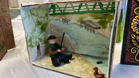 Tom Jackson/ BBC A wooden box has been painted to depict  a nature scene with a river, trees and grass. Two small sculptures are inside the box. One made to look like a person fishing in the painted river and another resembling a duck. A model bucket has also been made as well as a small fish on the end of the fishing rod.