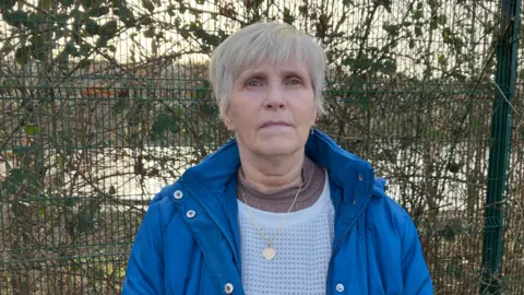 A woman stands in front of fencing. She wears a blue jacket, has short silver hair and is wearing a gold heart necklace