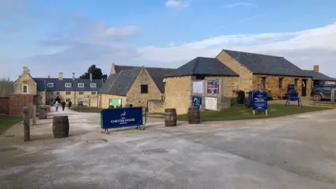 Martin Heath/BBC Chester House showing the original three-storey stone farmhouse to the left and other converted buildings housing various exhibits and experiences. There is a CHESTER HOUSE blue banner at the entrance, alongside barrels acting as vehicle barriers.