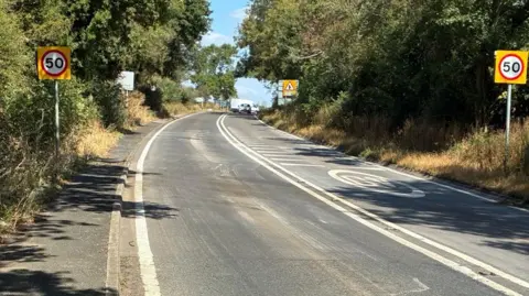An A road, with road markings. Trees line the road on either side side with 50mph speed limit signs either side.