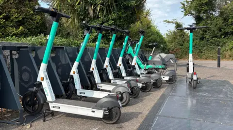 A row of e-scooters for rent at the side of a pavement on a sunny day in Cornwall. The vehicles have a white base and a turquoise steering column and black handlebars. They are lined up and secure by a black metal security rack. There are trees in the background.