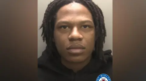 A man with black hair, wearing a black hoodie, stands in front of a plain wall in a police mugshot.