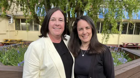 Headteacher Laura Mackie wearing a yellow blazer-style jacket and a black top underneath. She is smiling at the camera with brown, shoulder-length hair and is standing next to Helen who is wearing a black cardigan and gold necklace. She is smiling at the camera and has long brown hair. 
The two women are next to each other in front of a tree with bluebells beneath it and there is a building with lots of windows behind them. 