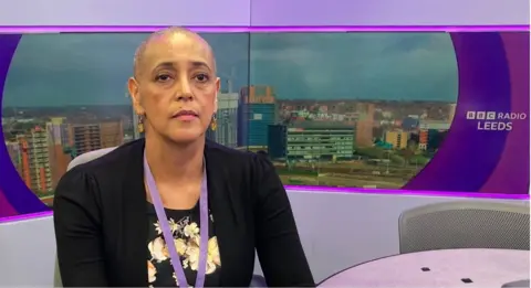 A woman with a shaved head wearing a black cardigan sitting in a radio studio.
