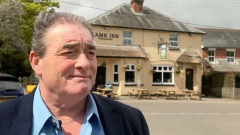 Duane is wearing a navy jacket and blue shirt and is standing across the road from his pub, the Lambs Inn, which is visible in the background.