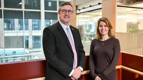 Councillor Michael Mordey and North East Mayor Kim McGuinness smiling next to each other at Sunderland City Hall. Michael Mordey is wearing a black suit with a white shirt and a pink tie. He has short grey hair and round glasses. Kim McGuinnes is wearing a brown dress and has long wavy blonde hair.