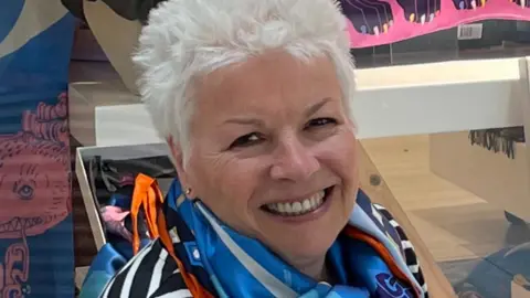 Francesca Tambling A woman with short white hair is wearing a striped top and a brightly coloured scarf, and smiling while seated indoors with patterned items displayed behind.