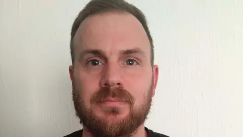 A headshot of Tony Pascall. He is standing in front of a white wall and looking directly into the camera.