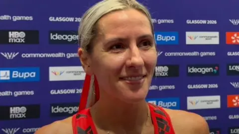 Manx Radio Christa Cain, who has long blonde hair tied back against the games board featuring partners and sponsors.