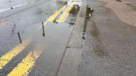 BBC A large puddle in the road over some double yellow lanes, with a pavement alongside.