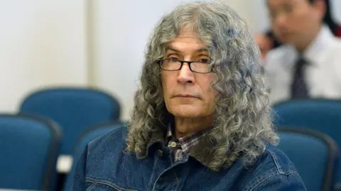 Getty Images Le tueur en série Rodney Alcala est assis en silence après avoir entendu la condamnation à mort prononcée par le juge Francisco Briseno dans une salle d'audience de Santa Ana le 30 mars 2010. Il porte une veste en jean, une chemise à carreaux et a de longs cheveux gris bouclés.