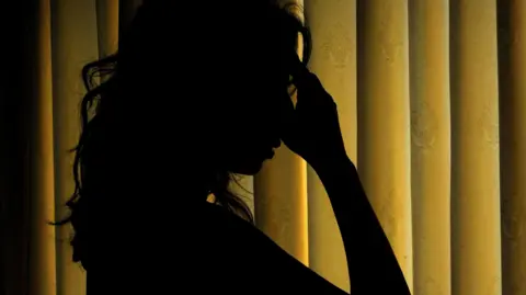 PA Media A woman with her head in hands. Only her silhouette is seen in a dark room with yellow curtains.