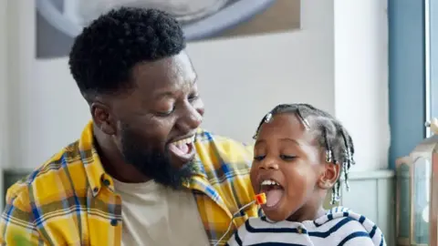 A man puts food in the mouth of a young child using a fork. The man is wearing a patterned yellow shirt and the child has braids and a blue stripy top on.