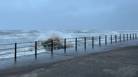BBC The Irish Sea seen from the promenade in Maryport. Tall waves are crashing on the promenade and the sky is grey with heavy clouds.